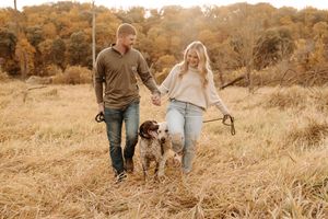 Couple walking hand in hand with spotted dog through golden autumn field.