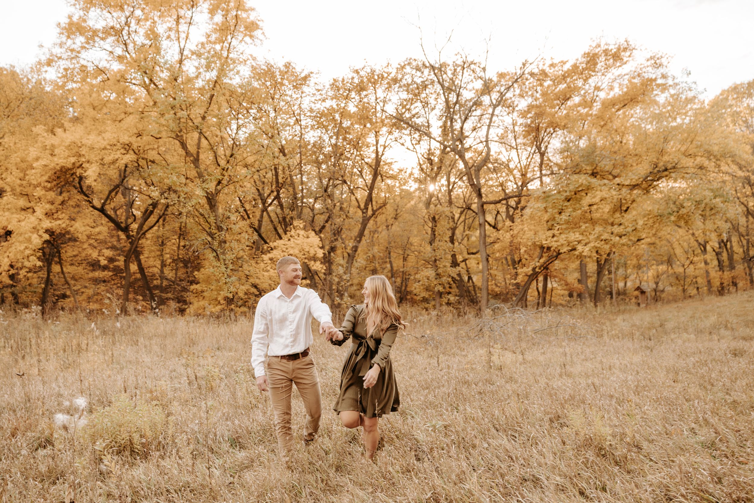 Couple holding hands walking through golden autumn field surrounded by vibrant fall foliage trees.