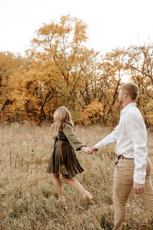 Couple holding hands in autumn field with golden trees, woman in olive dress, man in white shirt and tan pants.