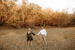 Couple holding hands in a golden autumn field surrounded by colorful fall foliage trees during an outdoor engagement photo session.