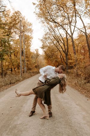 Couple sharing a romantic dip kiss on a dirt road surrounded by golden autumn trees in fall foliage.