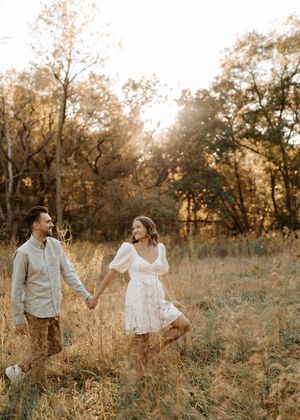 Couple holding hands walking through golden meadow at sunset, woman in white dress, man in casual attire.