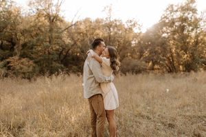 Couple embracing and kissing in a golden field surrounded by autumn trees during a romantic outdoor engagement session.