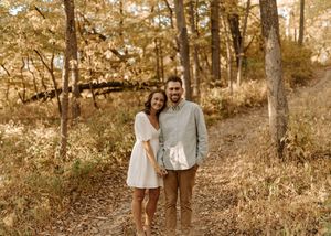 Couple smiling together on a forest path surrounded by autumn foliage and tall trees.