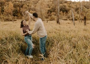 Couple dancing joyfully in a golden autumn field surrounded by tall grass and colorful fall trees.