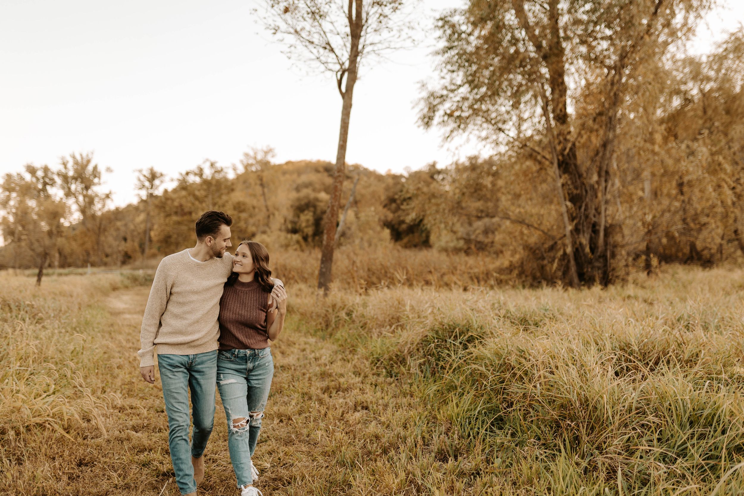 Couple walking together through an autumn field surrounded by golden trees and tall grass.