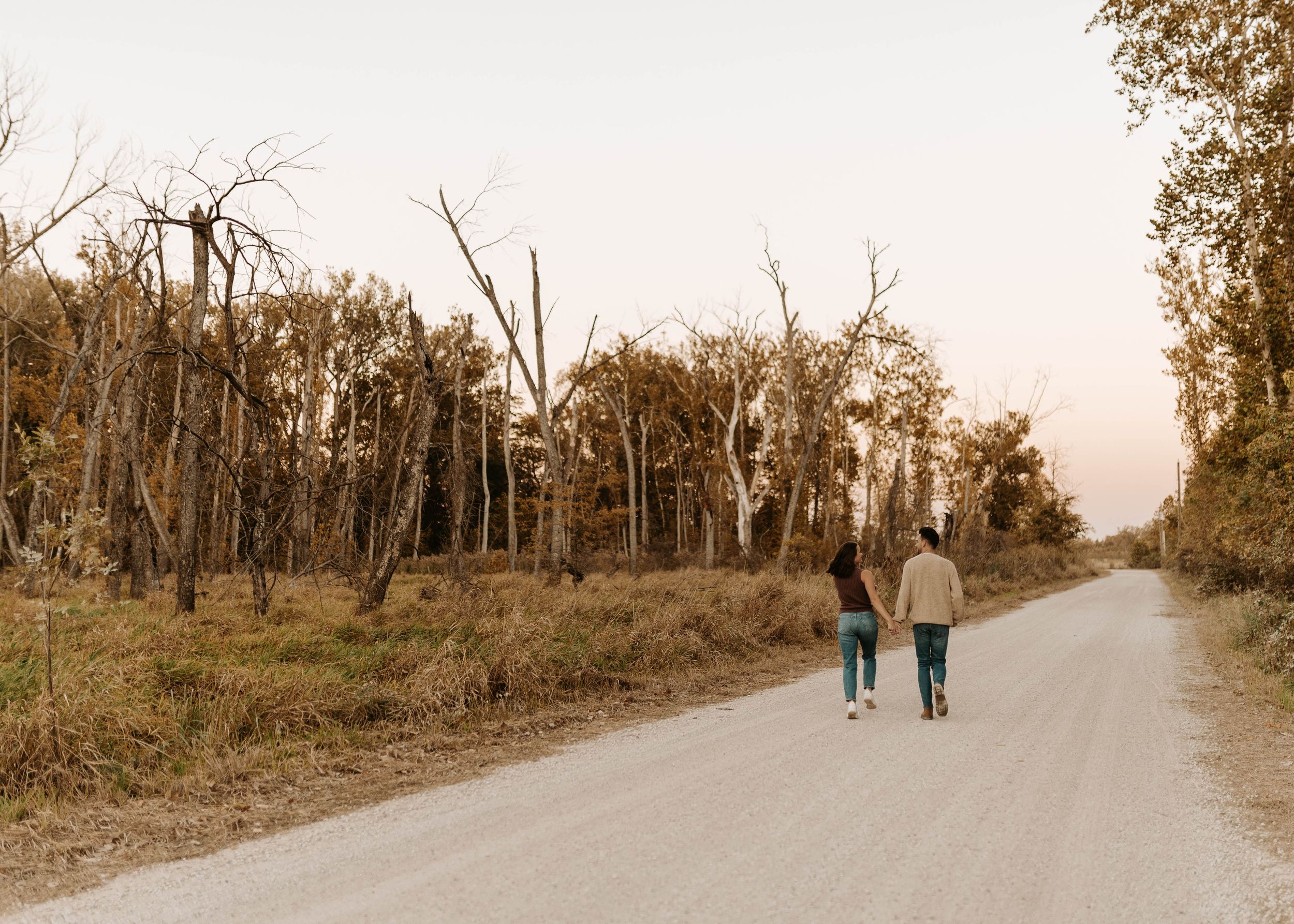 Two people walk hand in hand down a quiet gravel road surrounded by autumn trees at dusk.