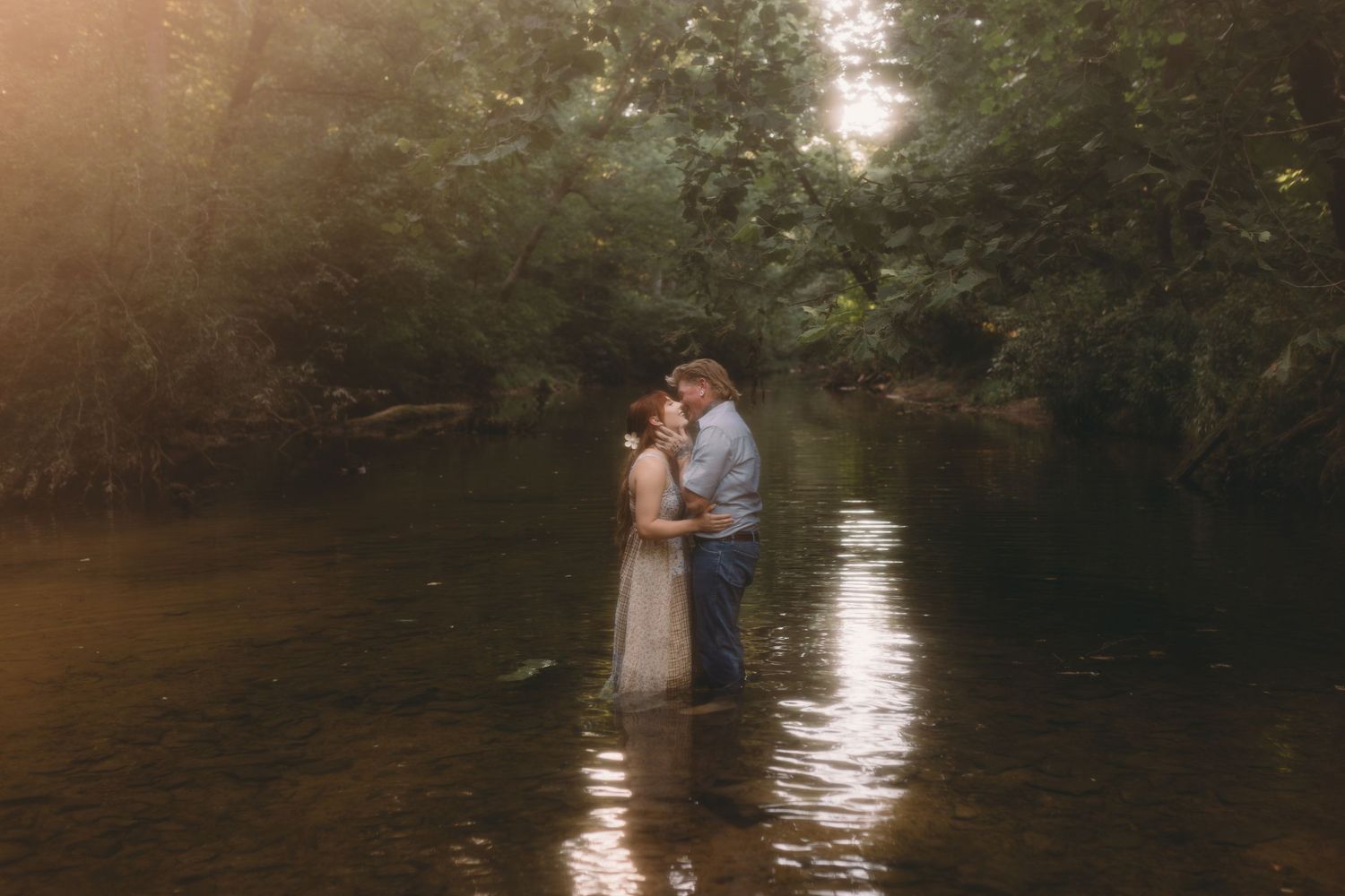 Couple standing together in a shallow creek surrounded by lush green trees in warm golden light.
