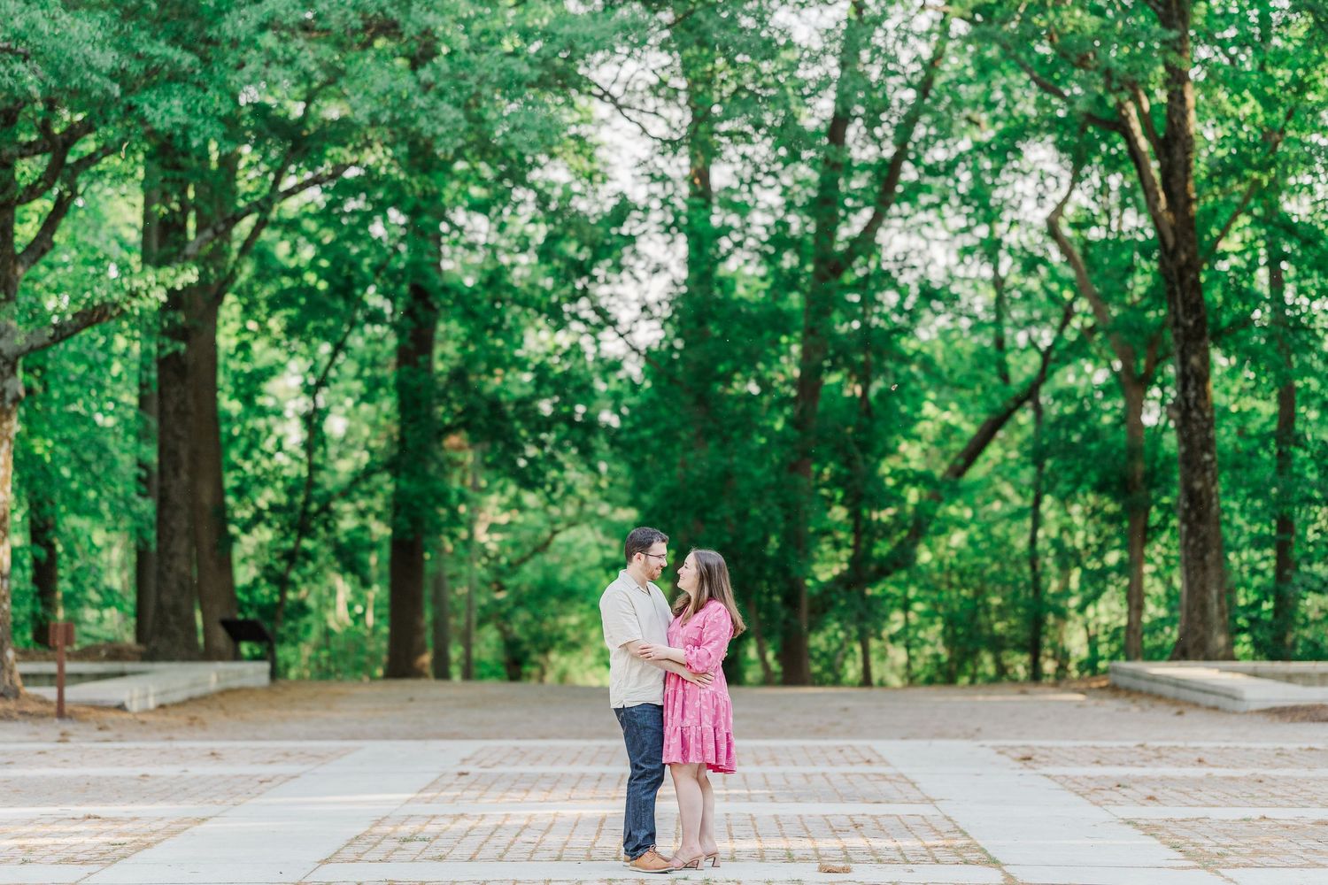 Couple embracing and smiling at each other on a brick path surrounded by lush green trees in a park.