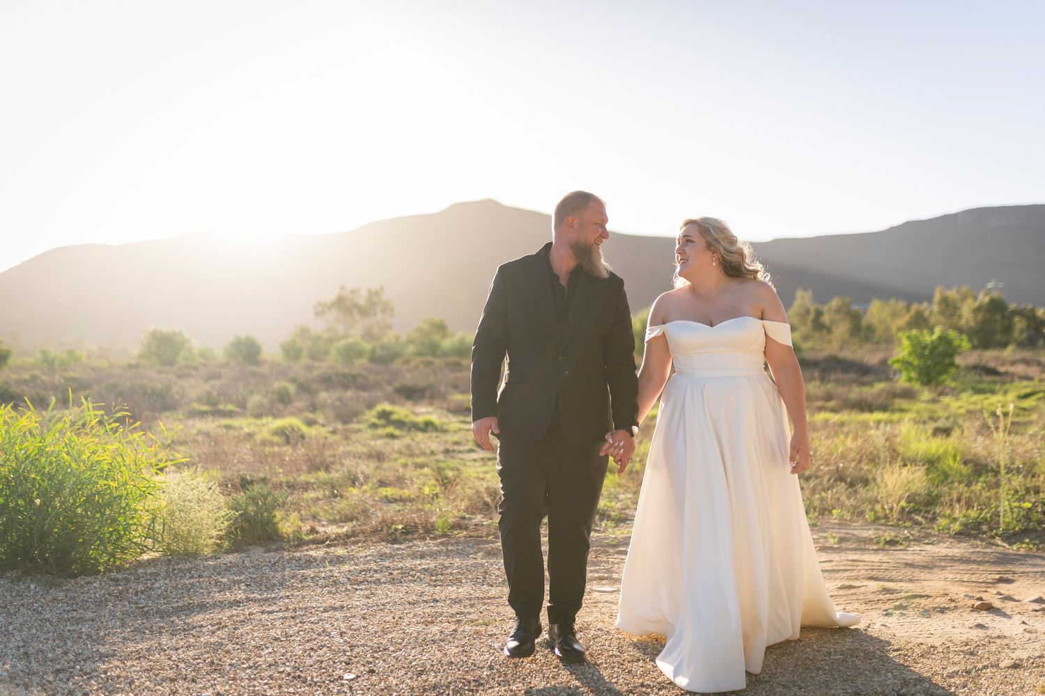 A couple in wedding attire holds hands walking through a desert landscape at golden hour with mountains in the background.