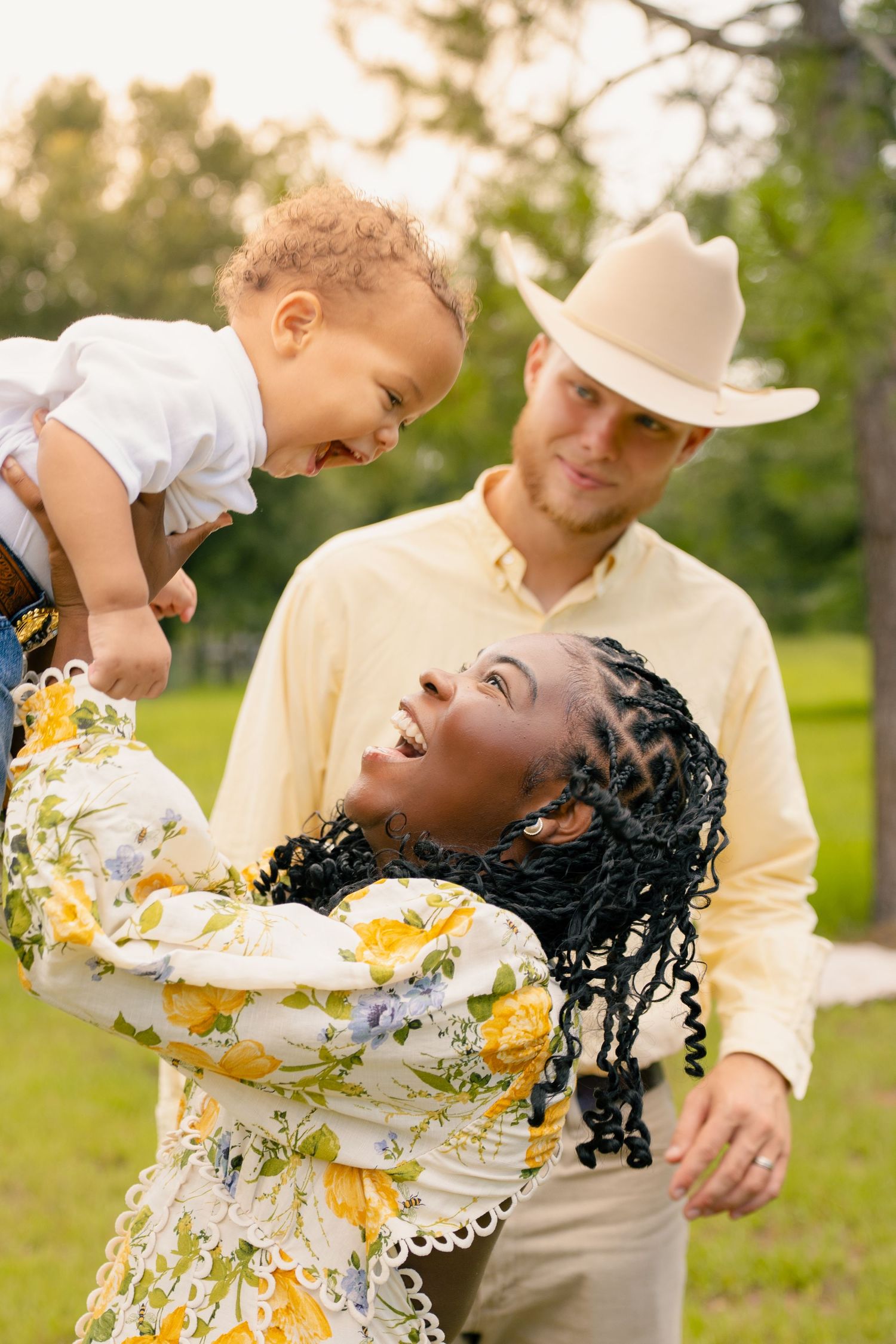 Happy interracial family playing outdoors, mother lifting laughing toddler while father watches in a sunny park.