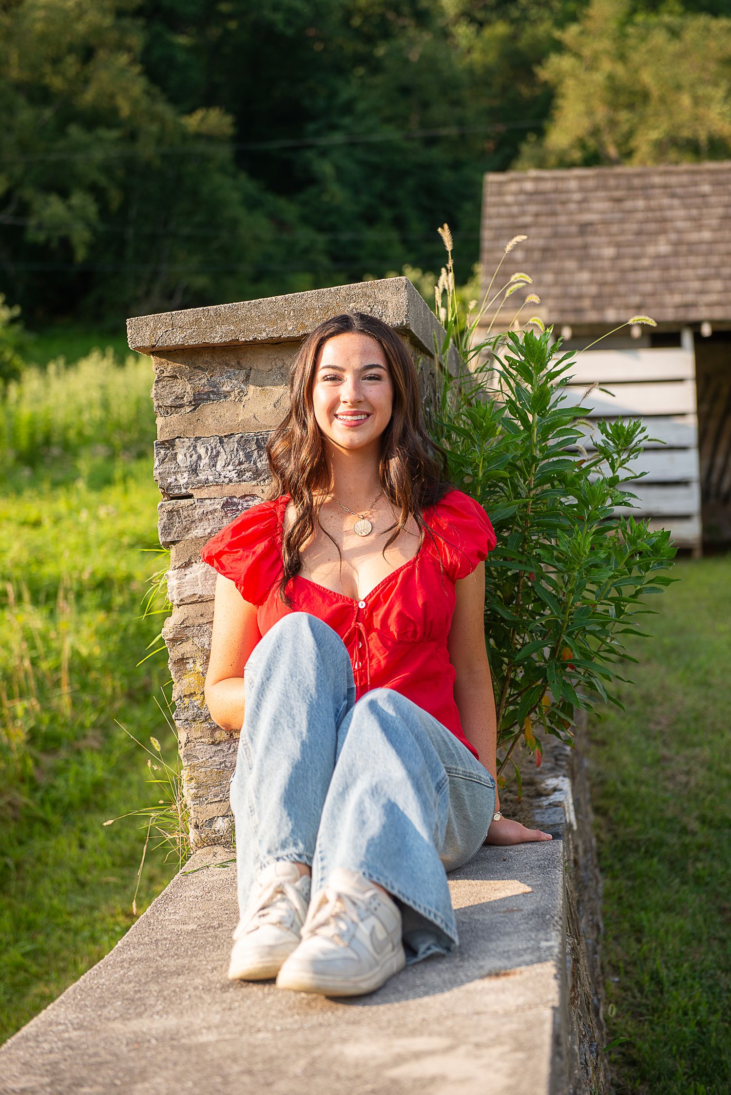 Young woman in red top and jeans sitting on stone wall outdoors, smiling, with green trees and rustic cabin behind her.