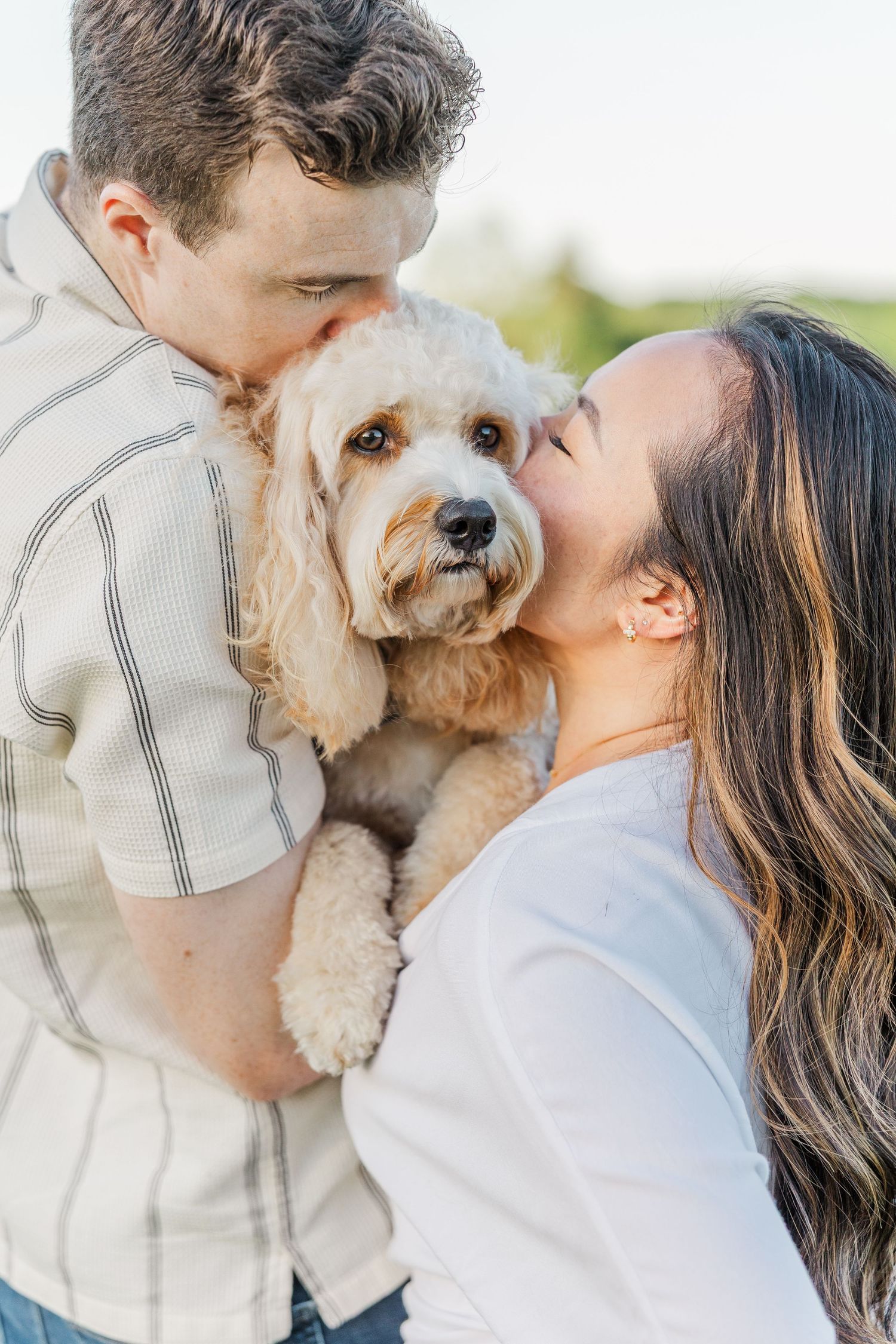 Couple kissing their fluffy golden doodle dog outdoors in a sweet engagement photo session.