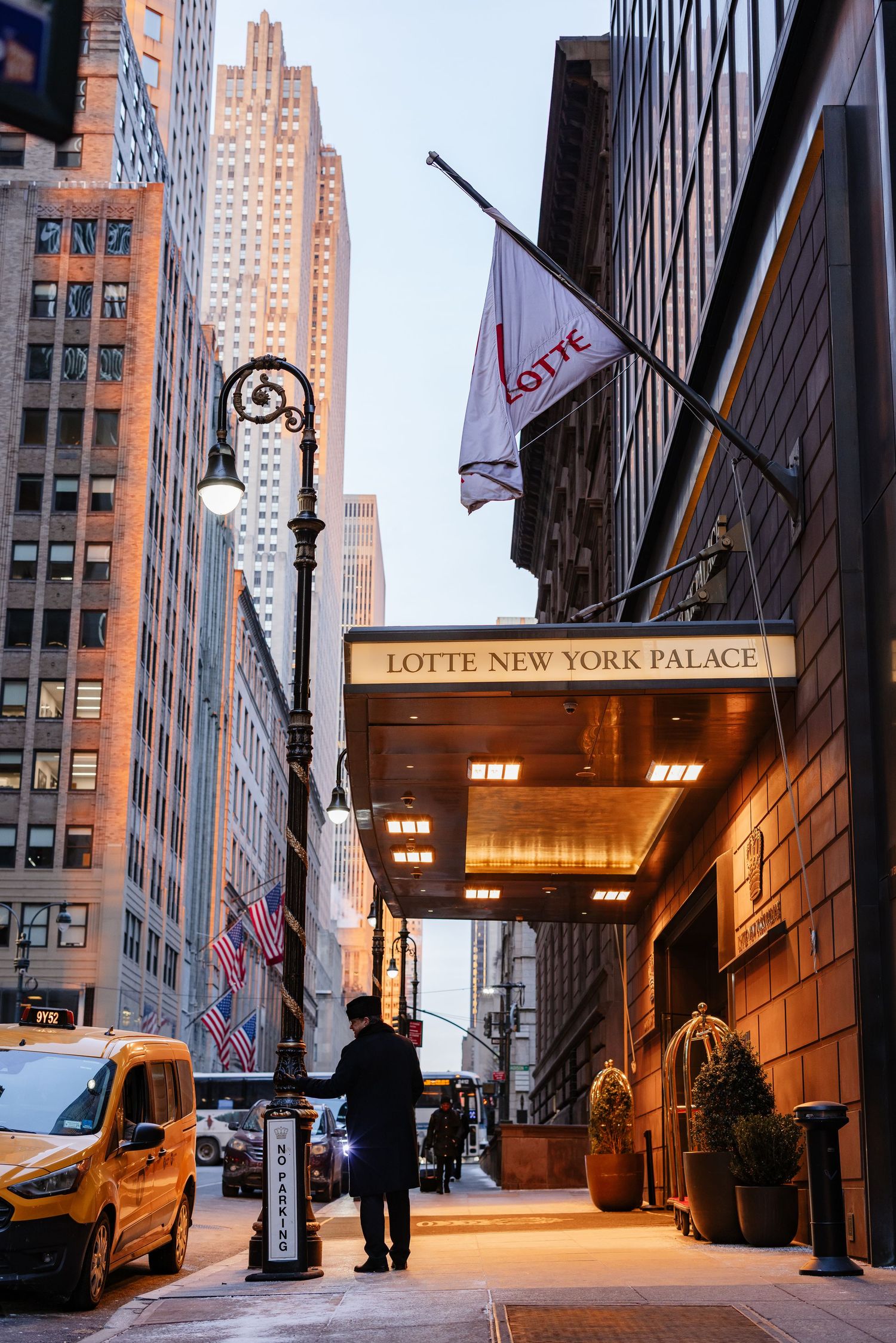 Person standing outside the Lotte New York Palace Hotel entrance with yellow taxis and skyscrapers in NYC.