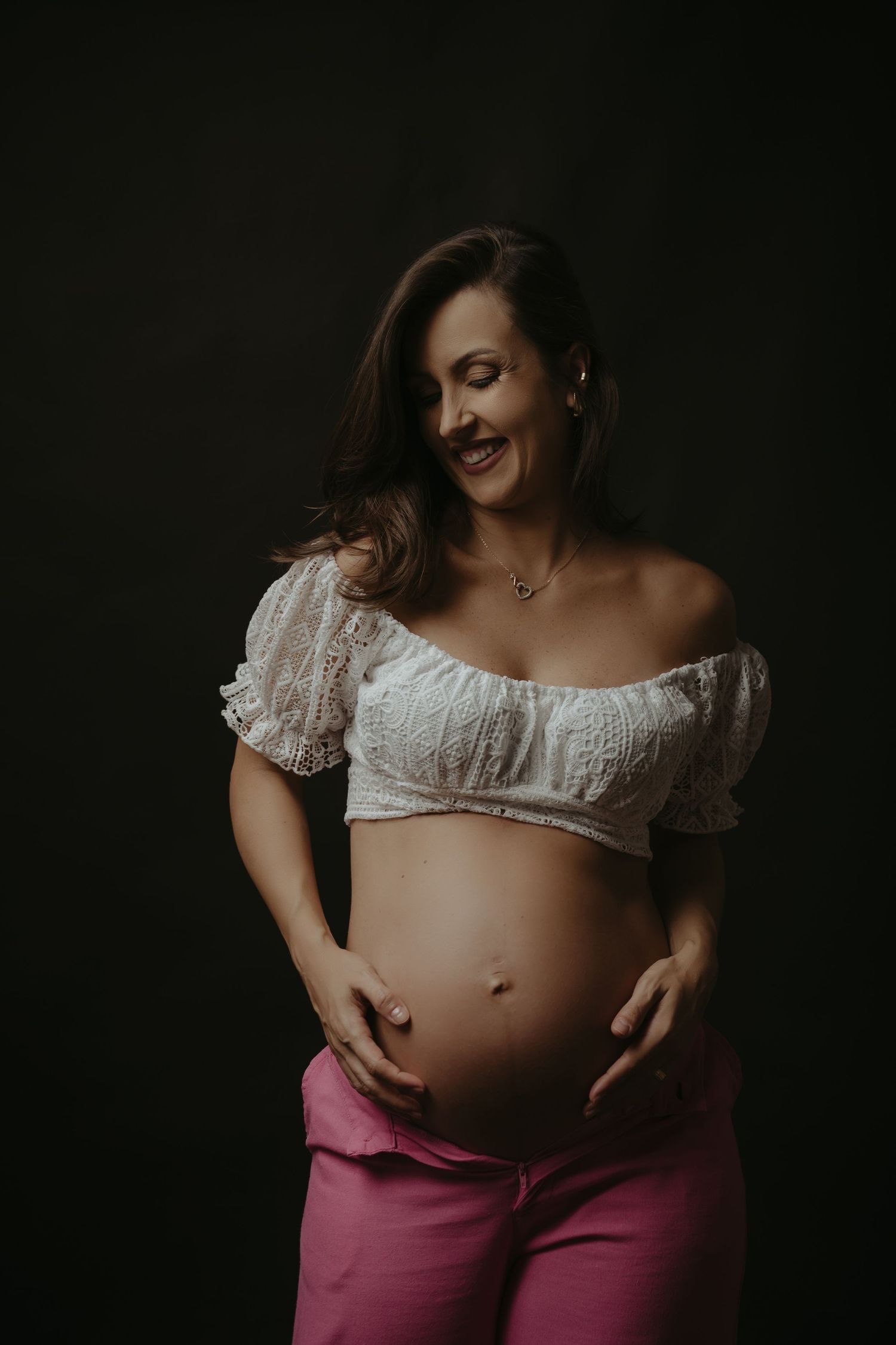 A pregnant couple in white shirts and jeans share a tender moment, smiling at each other while holding her baby bump.
