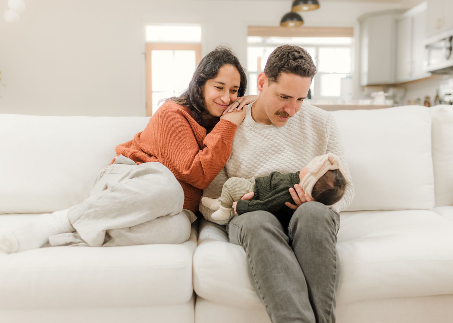A couple sits on a white sofa, smiling at their newborn baby in a bright, modern living room.