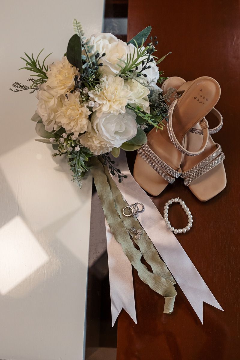 Bridal flat lay featuring white floral bouquet, nude heeled sandals, pearl bracelet, and wedding rings on wooden surface.