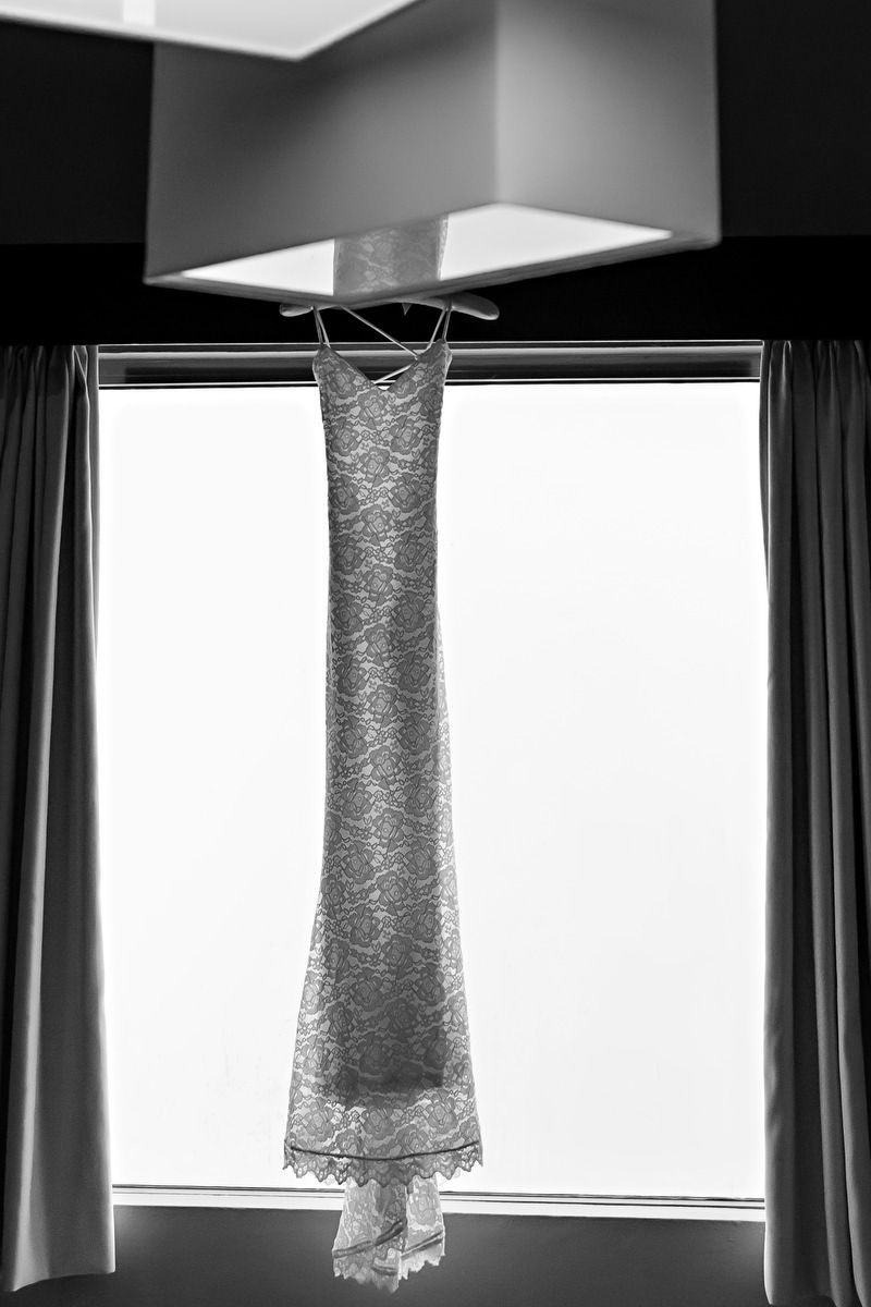Black and white photo of an elegant lace wedding dress hanging by a window, showcasing its slim silhouette.