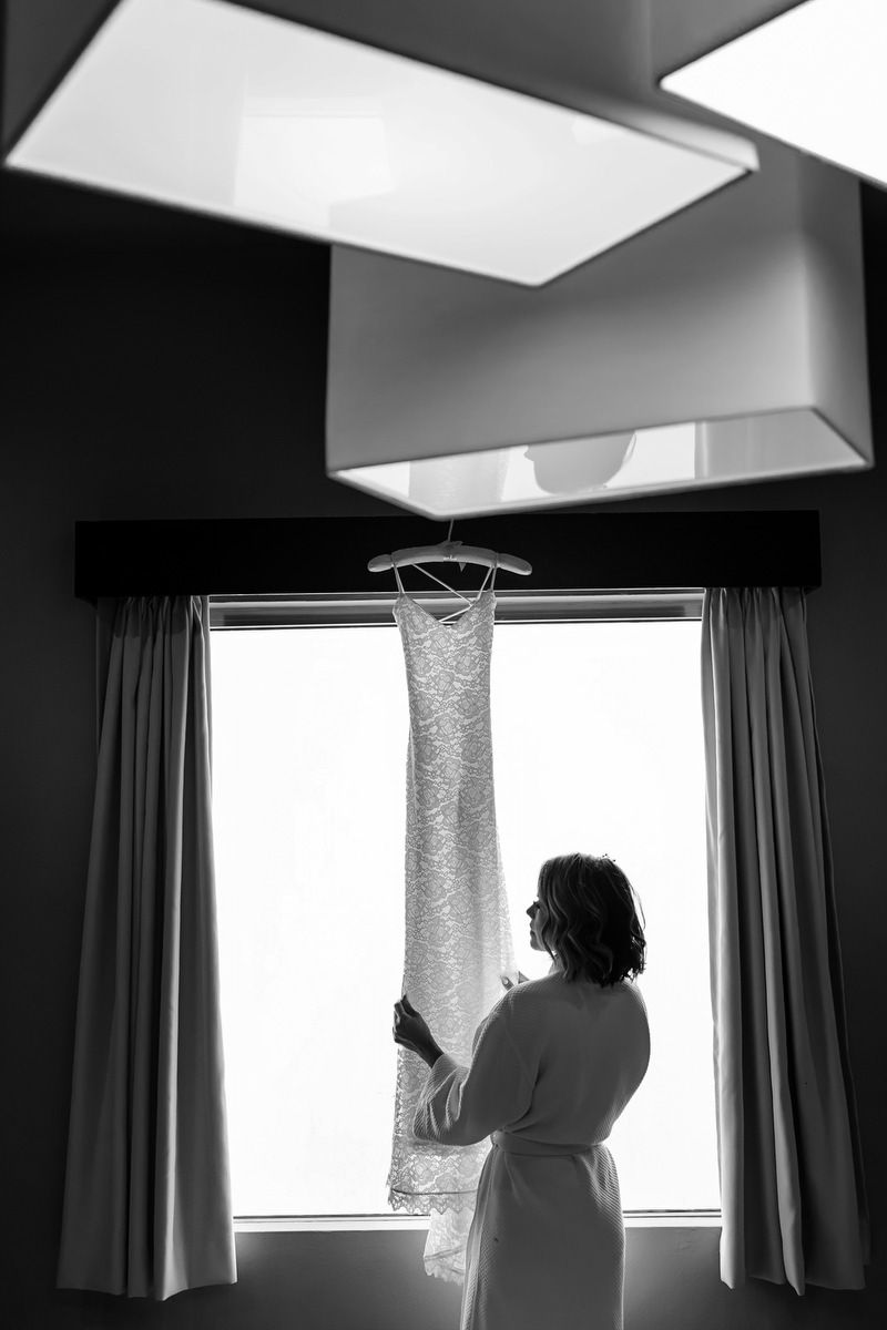 Bride in robe admiring wedding dress hanging by window in black and white bridal preparation photo.