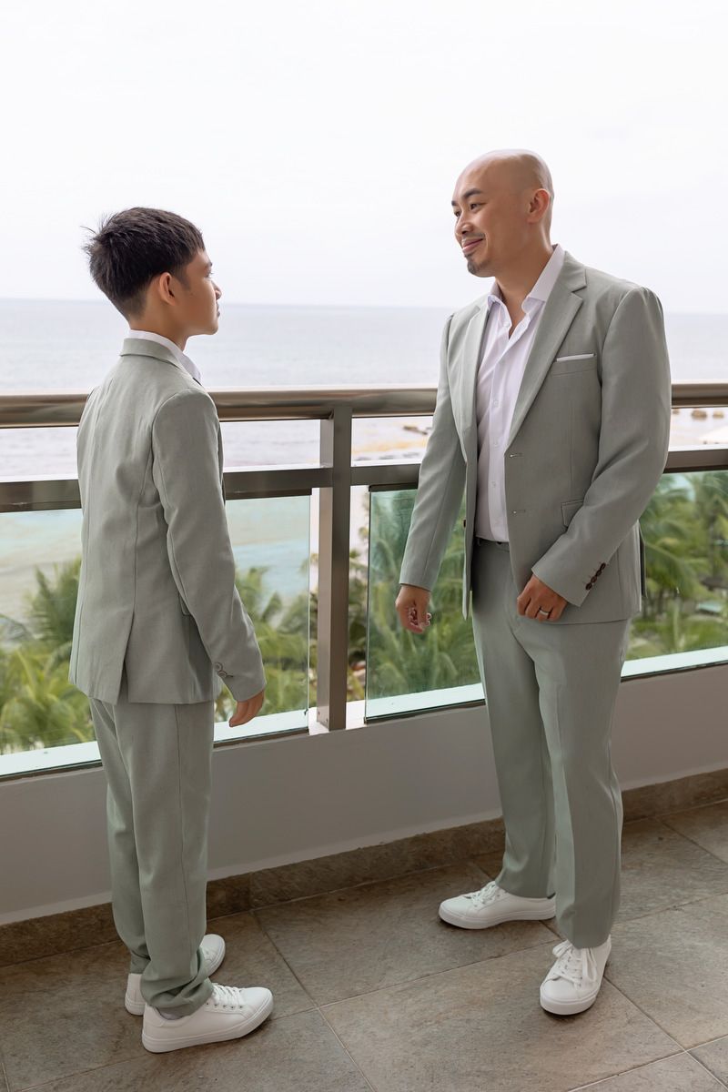 Two men in matching sage green suits stand on a balcony overlooking a tropical beach, smiling at each other.