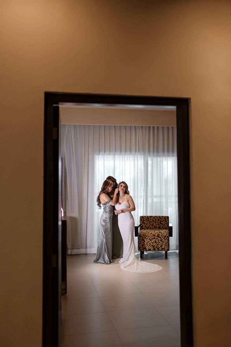 Bride in white gown being helped by bridesmaid in silver dress in elegant hotel room before wedding ceremony.