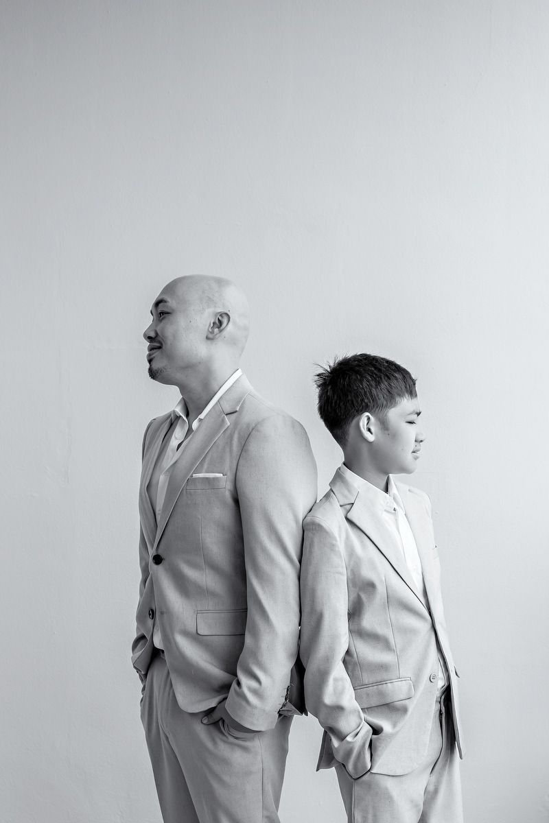 Black and white photo of a bald man and young boy in matching suits standing back to back.