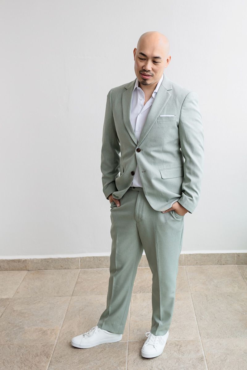 Bald man in sage green suit with white sneakers posing confidently against a light gray wall.