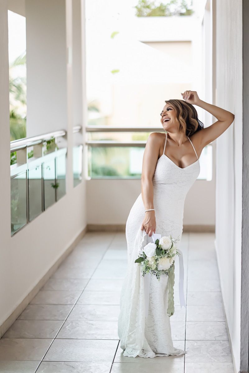 Joyful bride in white lace gown holding bouquet of white roses laughing on a sunlit balcony.