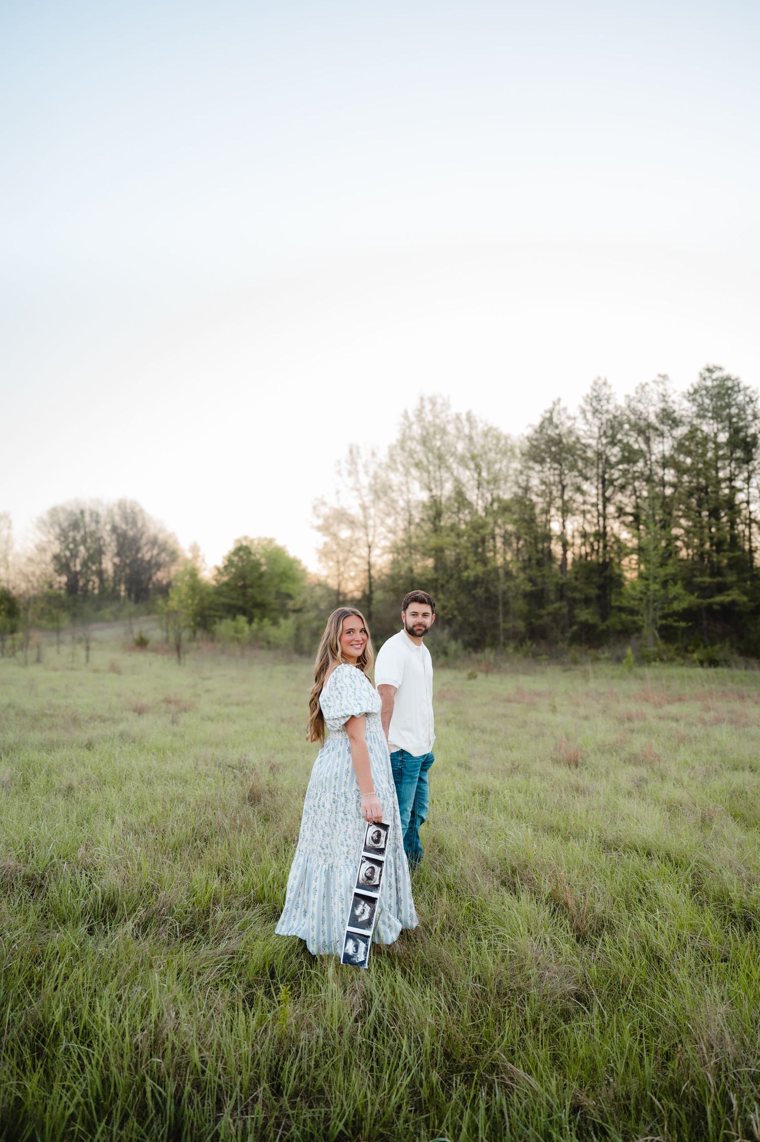 Couple standing back-to-back in a green meadow, woman in white dress holding ultrasound photos, announcing pregnancy outdoors.