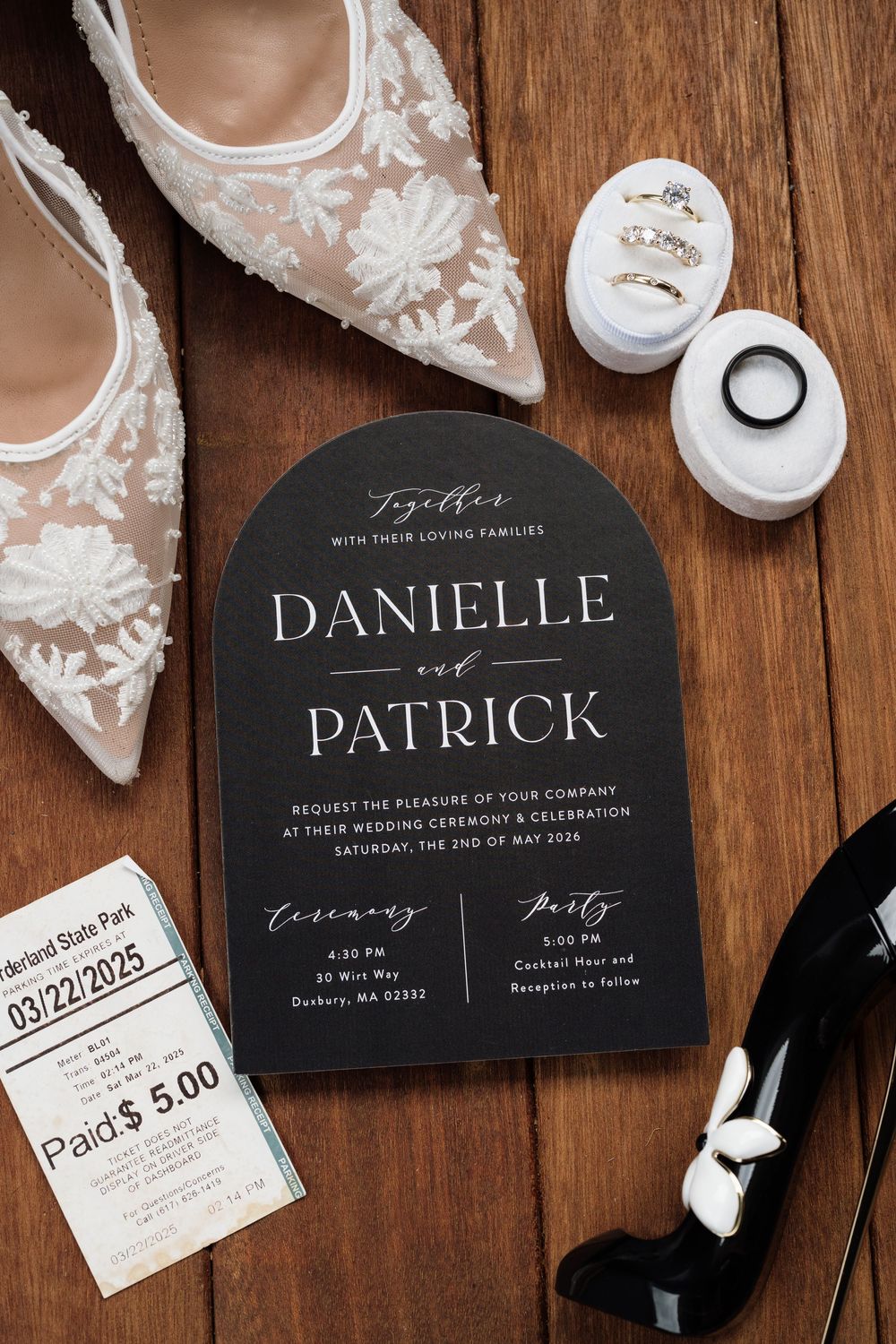 Elegant black arched wedding invitation for Danielle and Patrick on a wooden surface with lace heels and rings.
