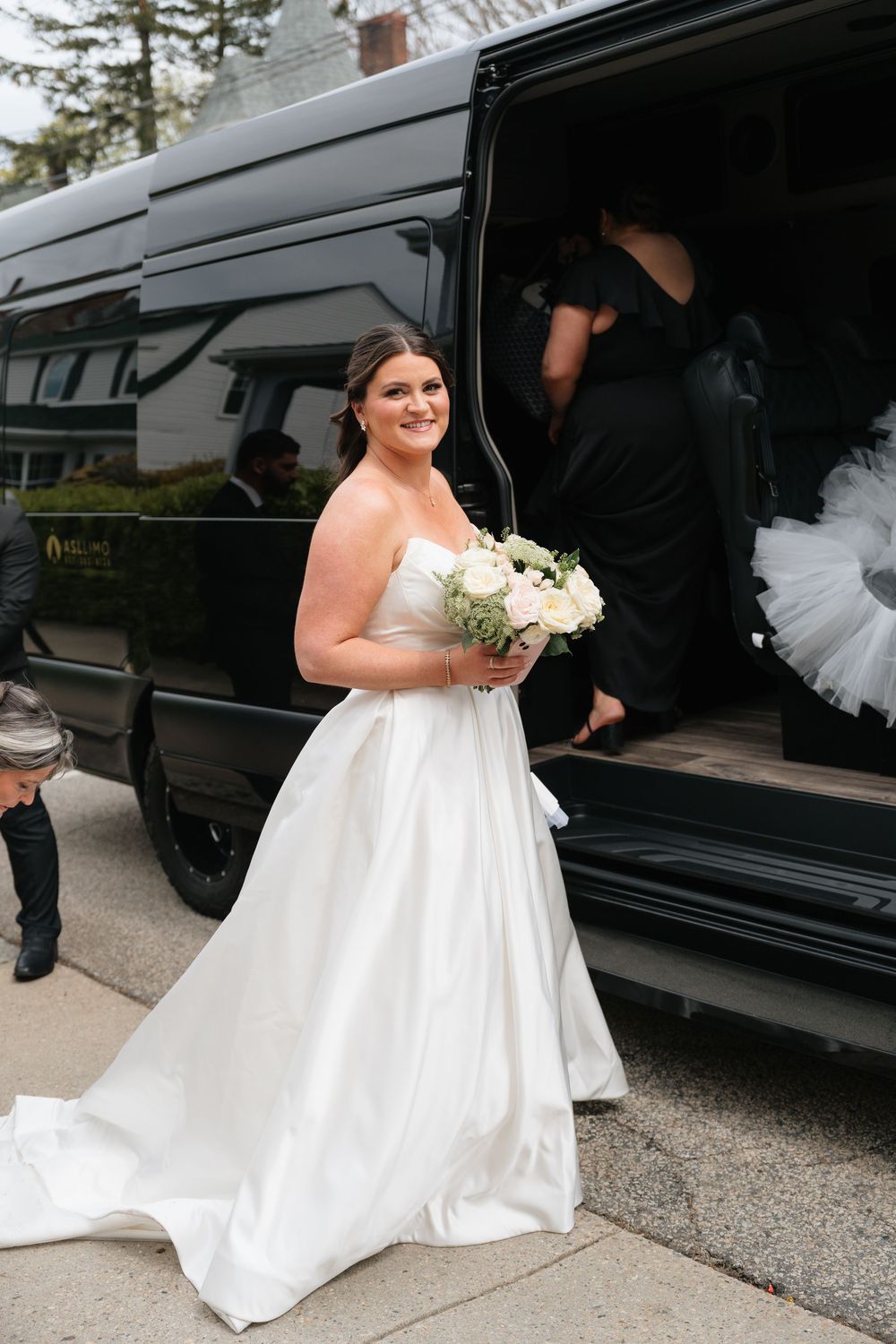 Smiling bride in strapless white ballgown holding bouquet stepping out of black wedding party van.