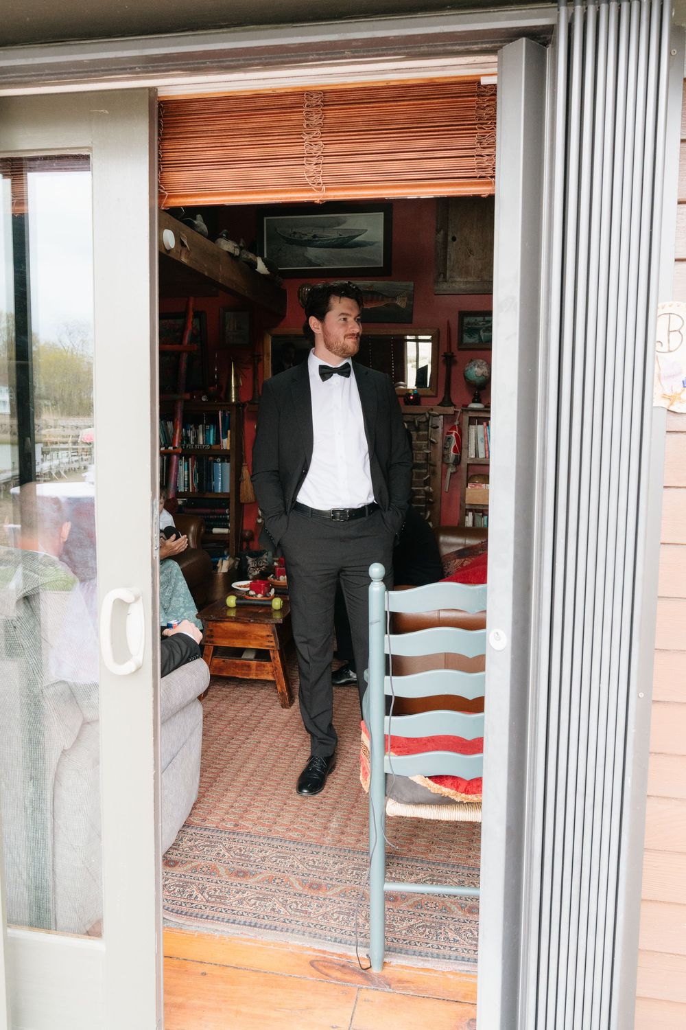 Groom in black tuxedo with bow tie standing in doorway of cozy library room before wedding ceremony.
