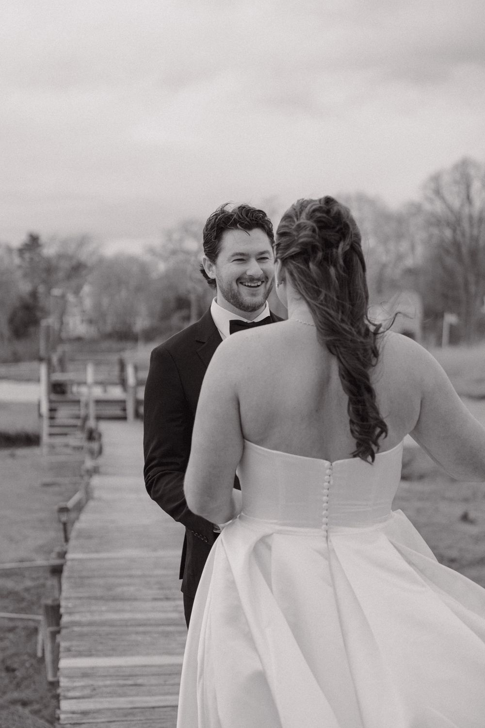 Black and white photo of a groom smiling joyfully at his bride on a wooden dock during their wedding day.
