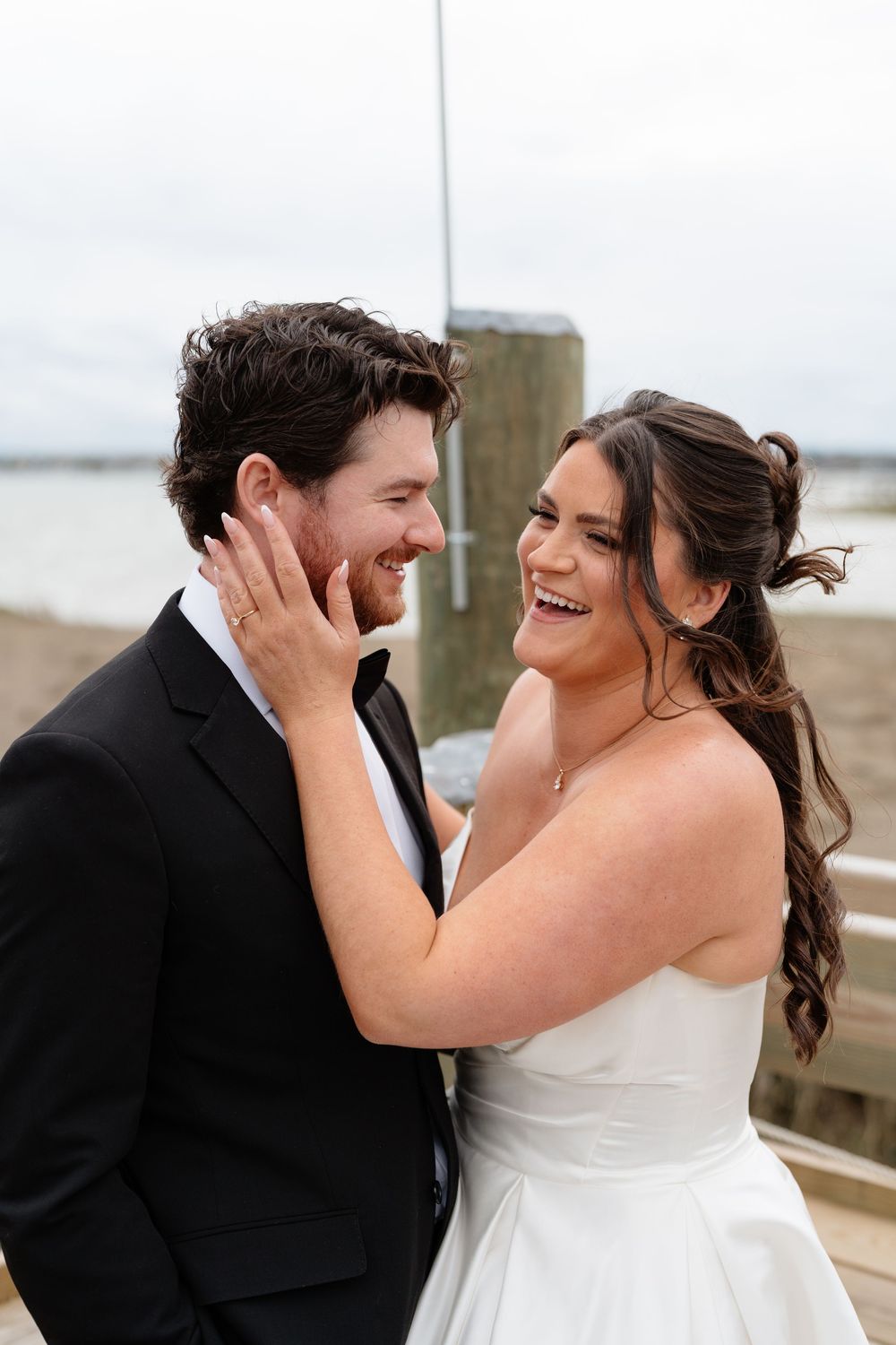 Happy bride and groom share a joyful moment laughing together on a waterfront dock on their wedding day.