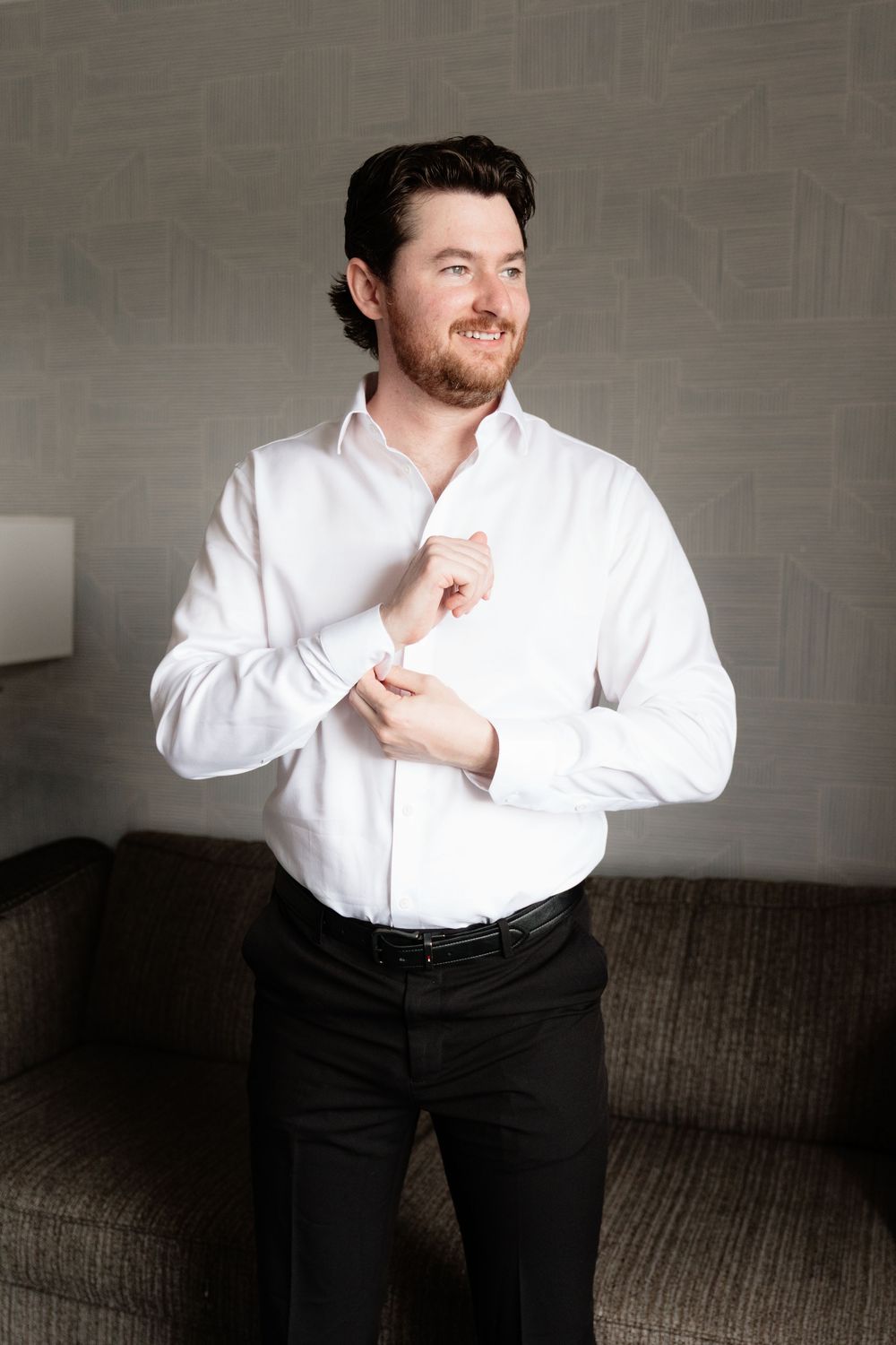 Groom in white dress shirt fastening cufflinks while smiling, preparing for wedding day.