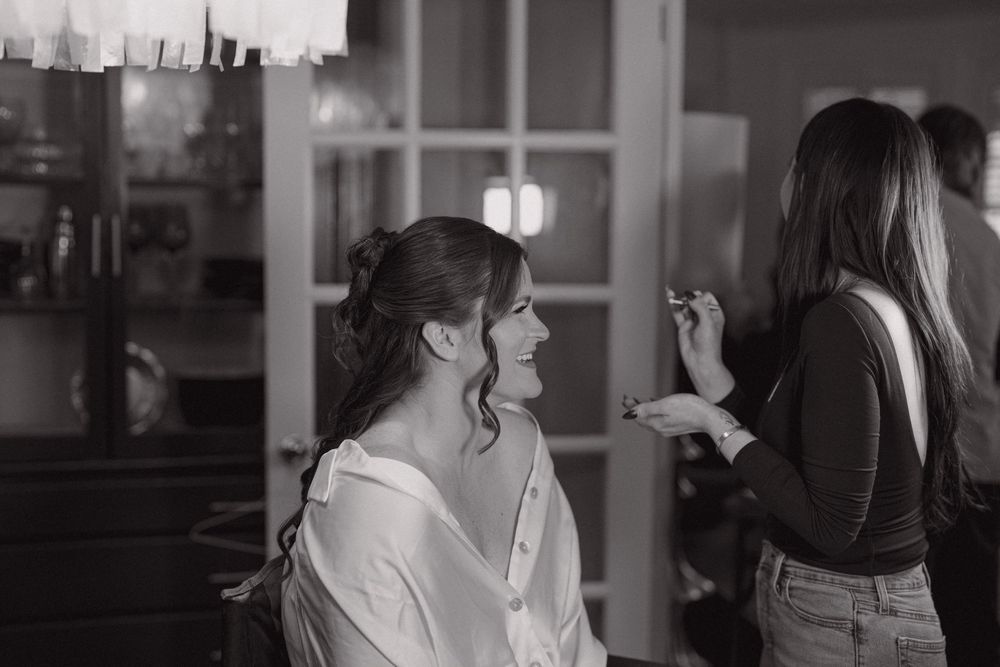 Bride in white robe smiles as makeup artist applies cosmetics in black and white photo.
