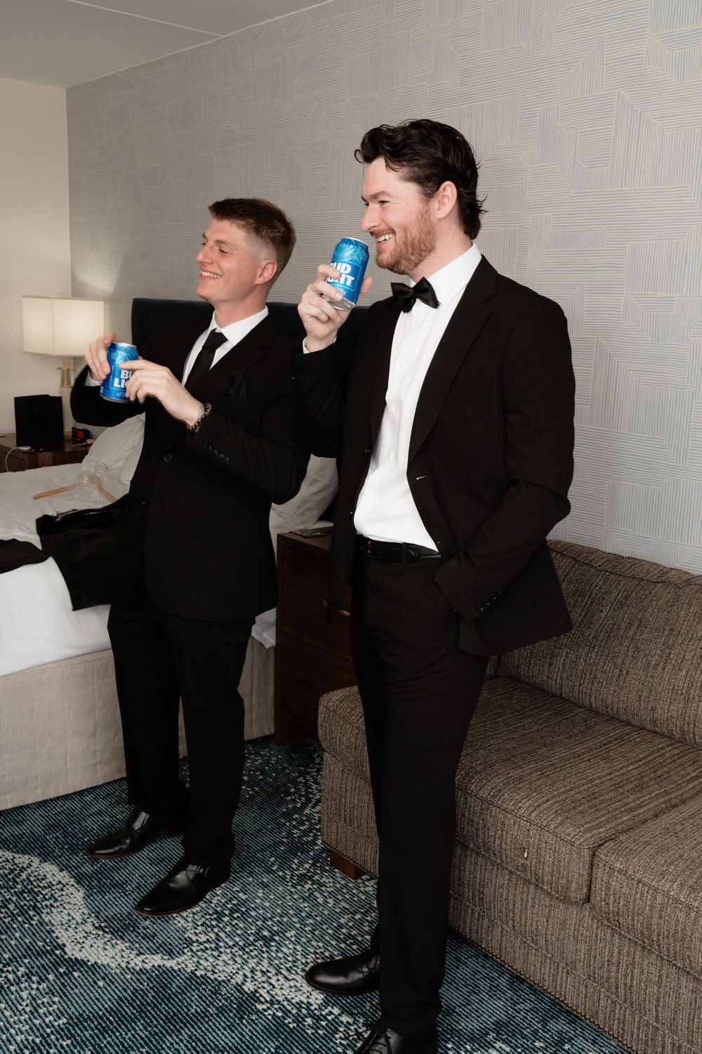 Two grooms in black tuxedos toast with Pabst Blue Ribbon beer cans in a hotel room before their wedding.