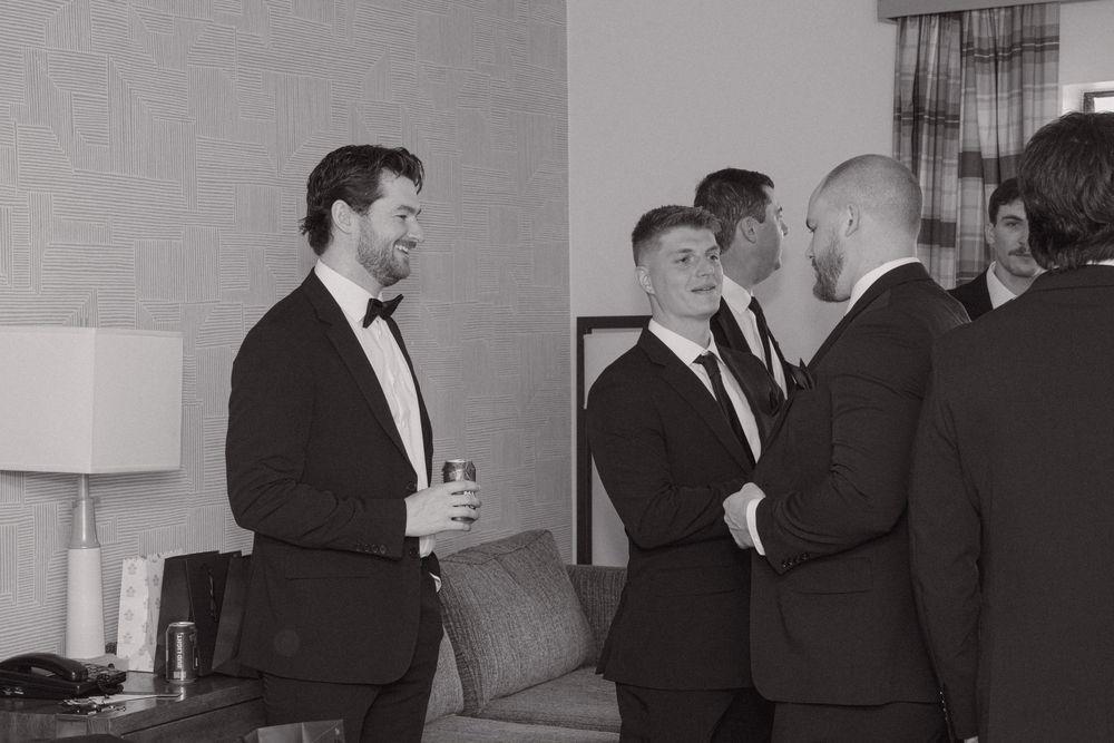 Black and white photo of groomsmen in tuxedos chatting and laughing together in a hotel room before a wedding.