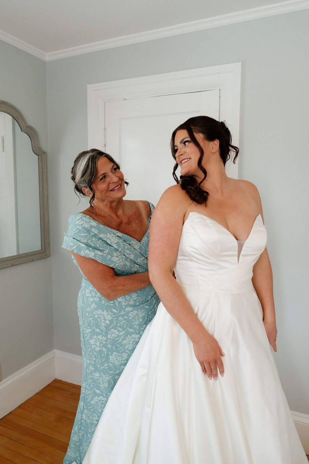 A bride in a white strapless gown smiles as an older woman in a blue floral dress helps her prepare for her wedding day.