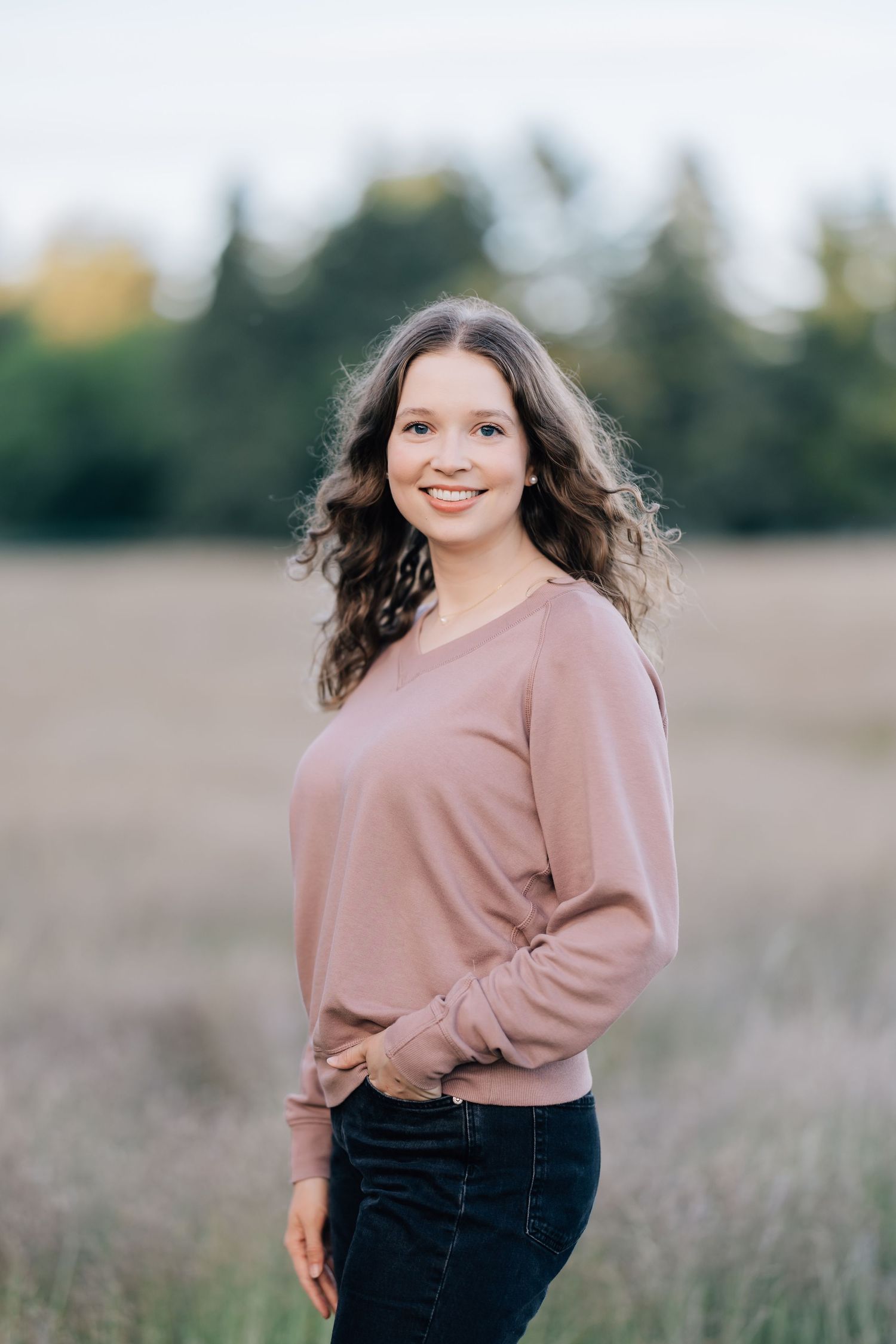 Young woman with curly brown hair smiling in a field, wearing a pink sweater and dark jeans with trees in background.