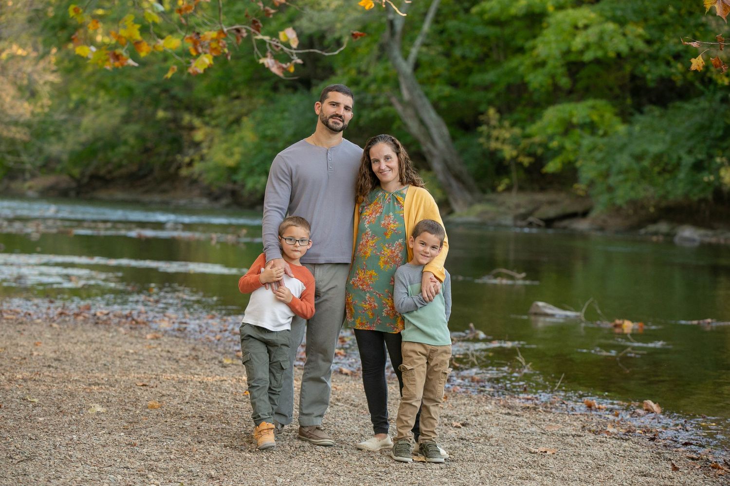 Family of four posing by a peaceful river surrounded by autumn foliage during an outdoor fall photo session.
