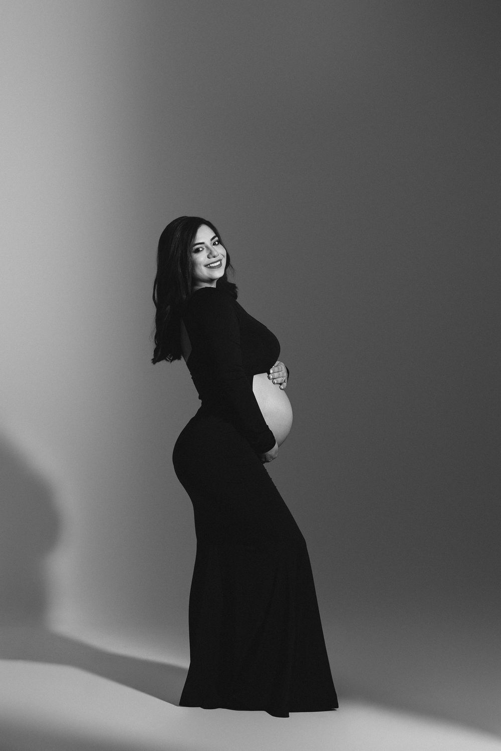 Black and white maternity photo of smiling pregnant woman in elegant black gown posing in studio.