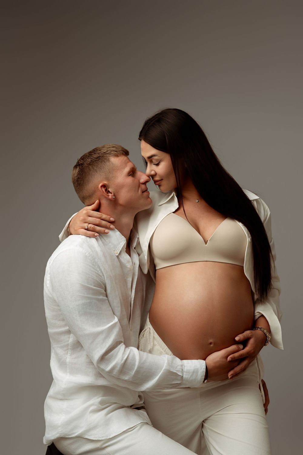 A couple in white clothing sharing a tender moment, with the man embracing his pregnant partner's belly in a studio.
