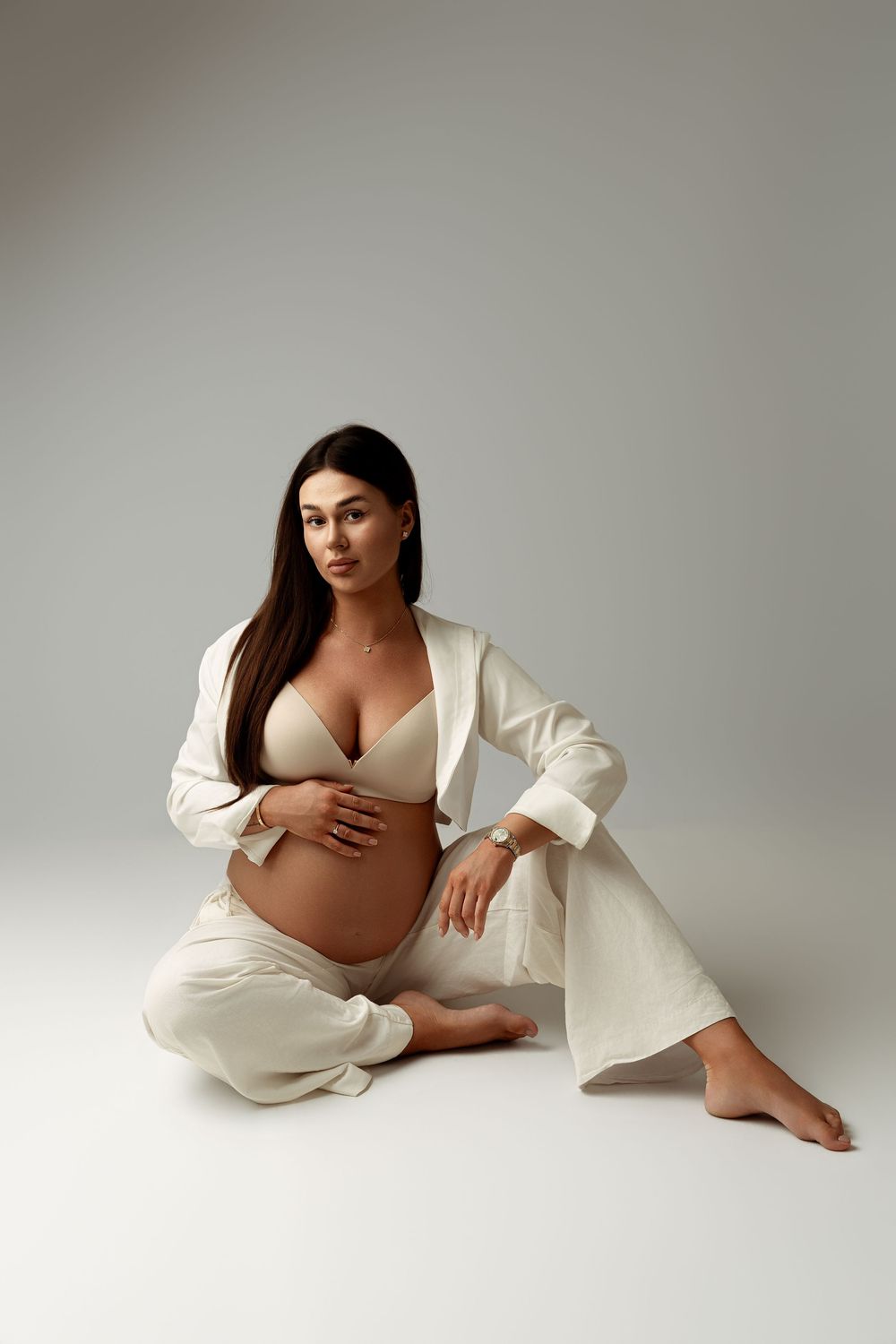 Pregnant woman in beige lingerie and white blazer sitting on floor cradling baby bump in studio setting.