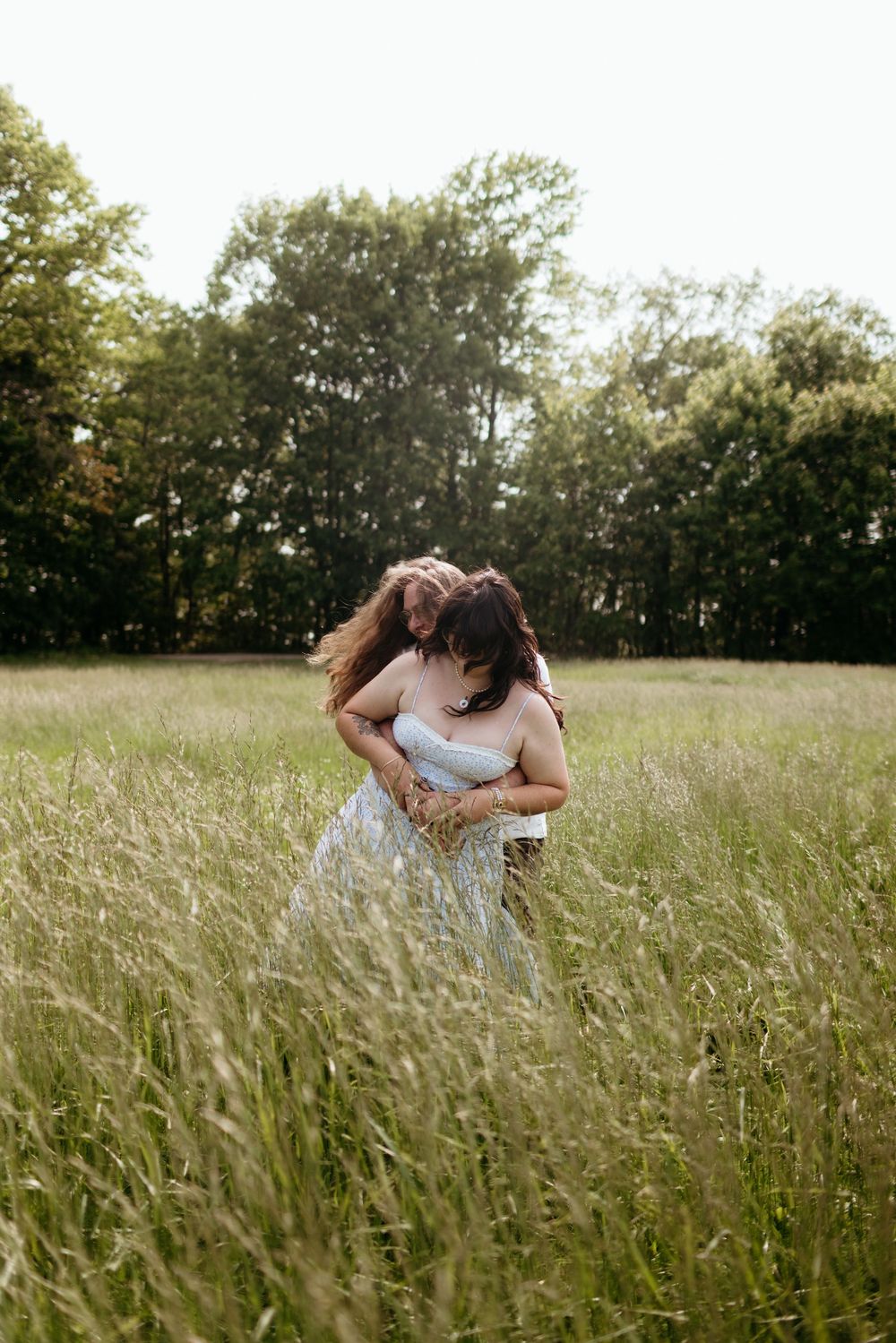 Two women embrace in a lush green meadow, wearing a white dress, surrounded by tall grass and trees.
