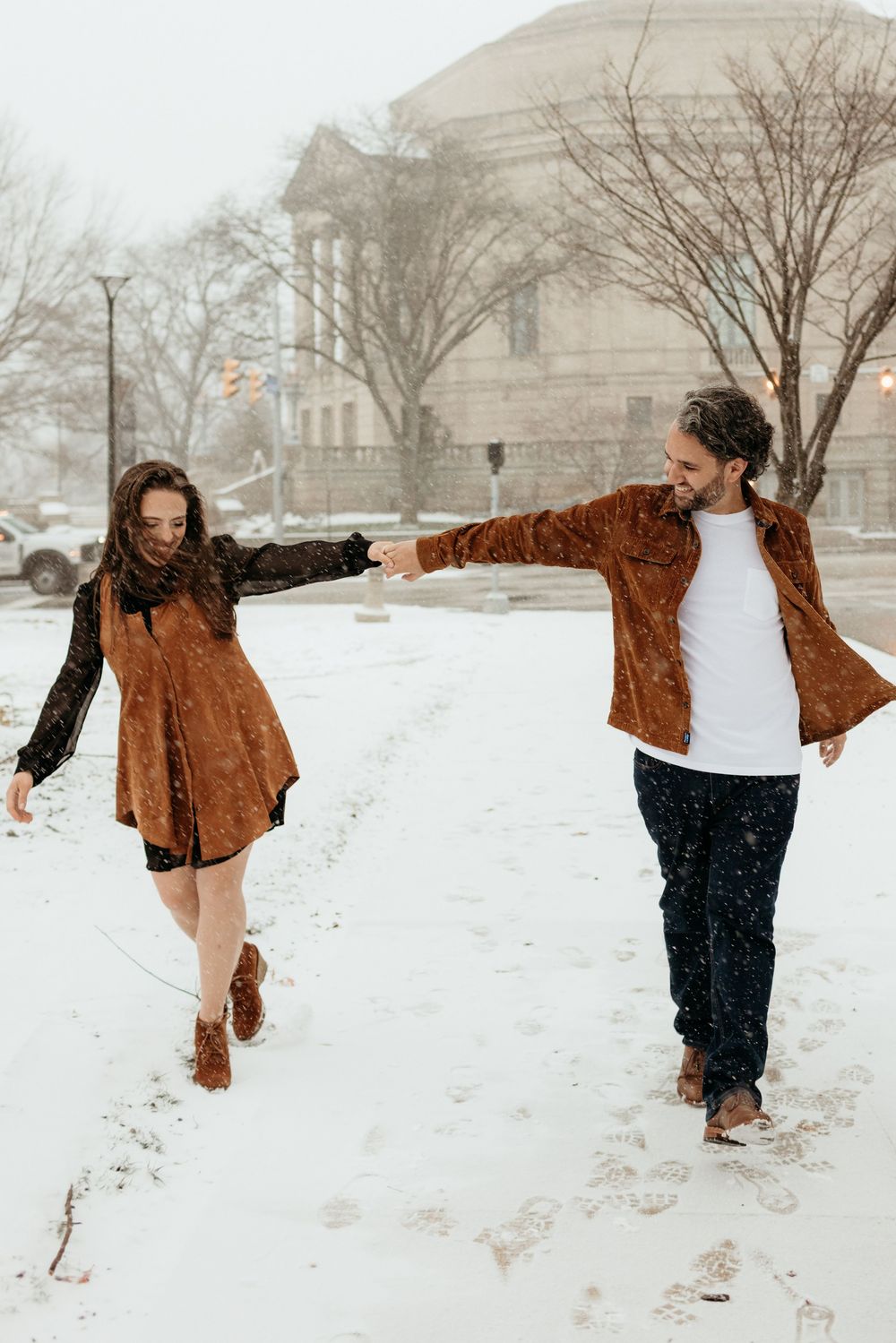 Couple holding hands walking in snow wearing matching brown suede outfits during a romantic winter engagement photo session.