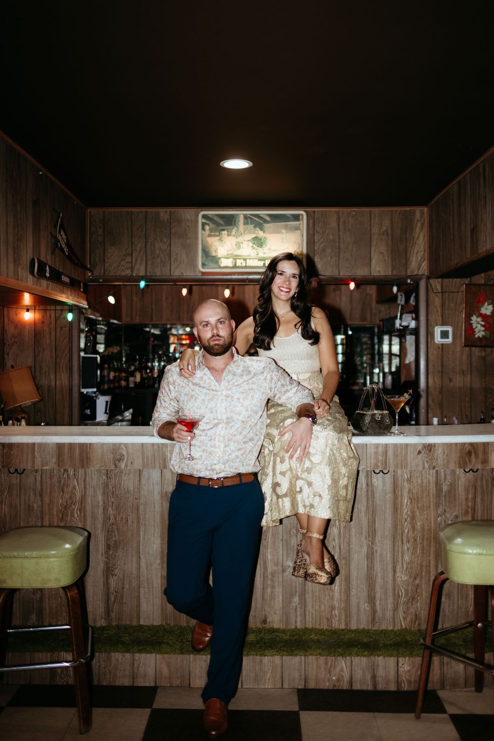 Couple posing at a rustic wooden bar, man holding cocktail in floral shirt, woman in gold dress seated on bar.