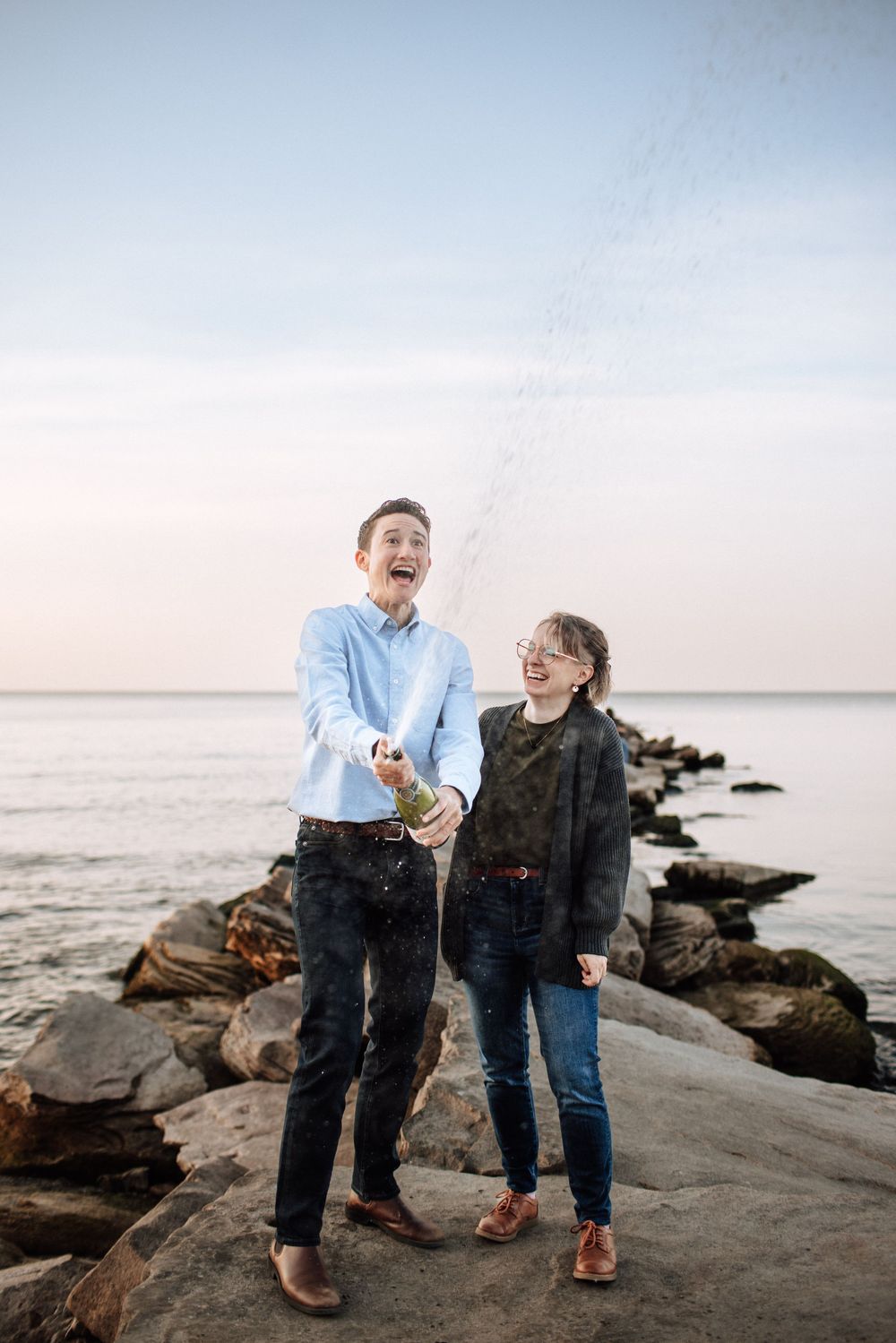 Couple laughing and popping champagne bottle on rocky shoreline at sunset, celebrating outdoors by the ocean.