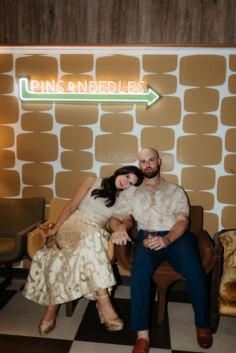 Couple posing together under a neon Pins & Needles sign on retro chairs with gold patterned wallpaper.