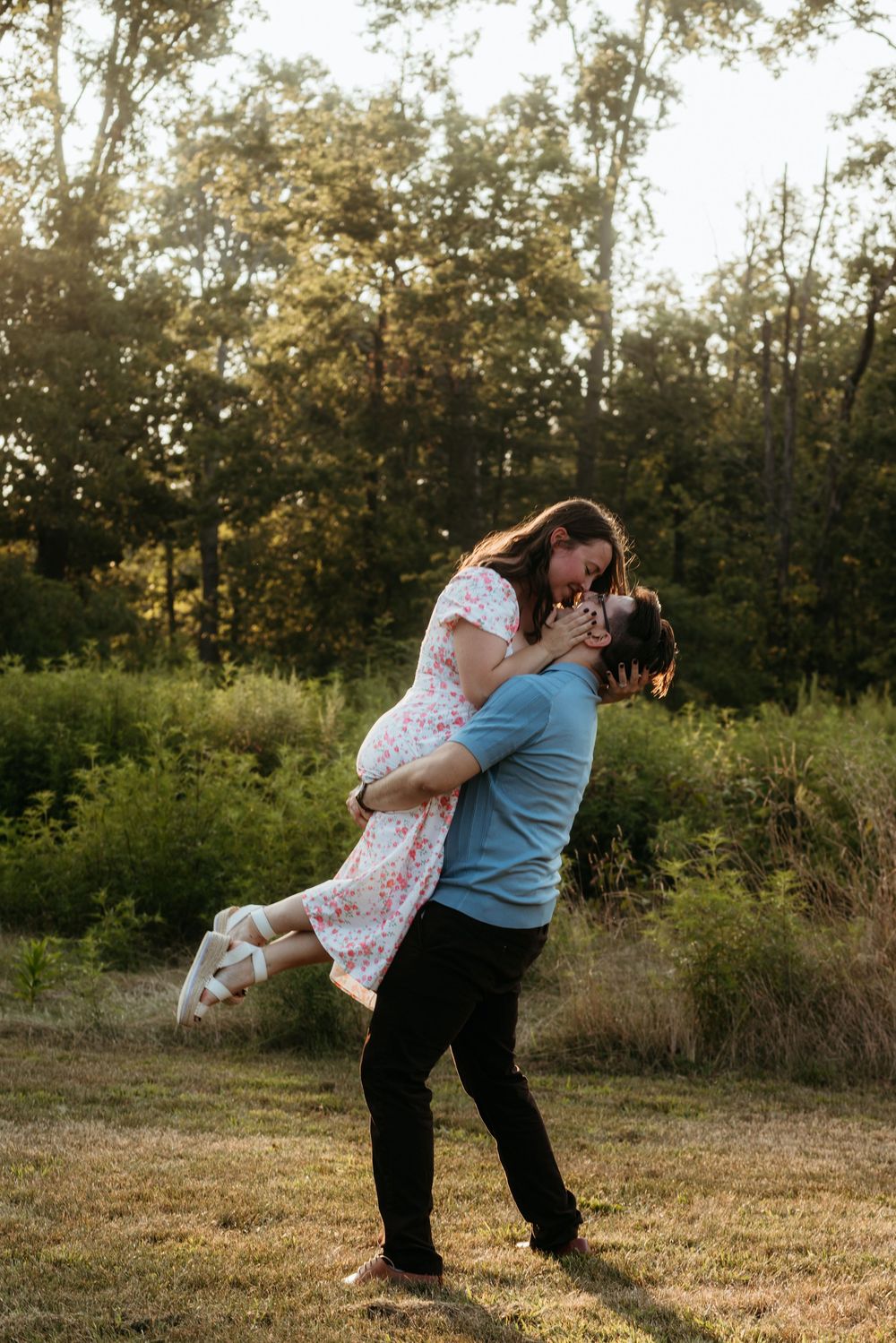 A man lifts a woman in a floral dress outdoors, sharing a romantic moment surrounded by lush green trees.